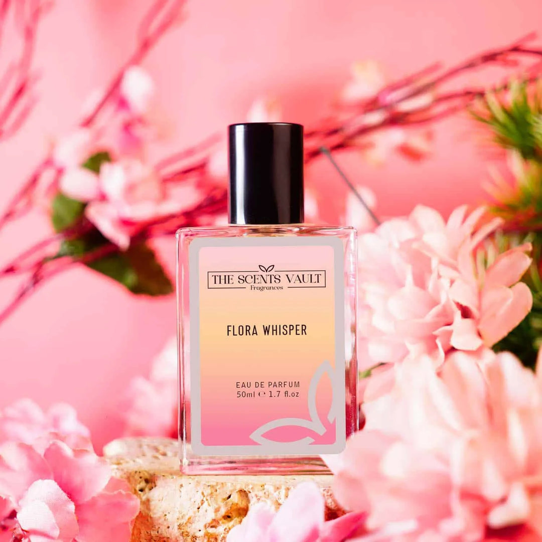 A bottle of 'Flora Whisper' perfume from The Scents Vault, eau de parfum, with a pink label, displayed against a backdrop with pink flowers.